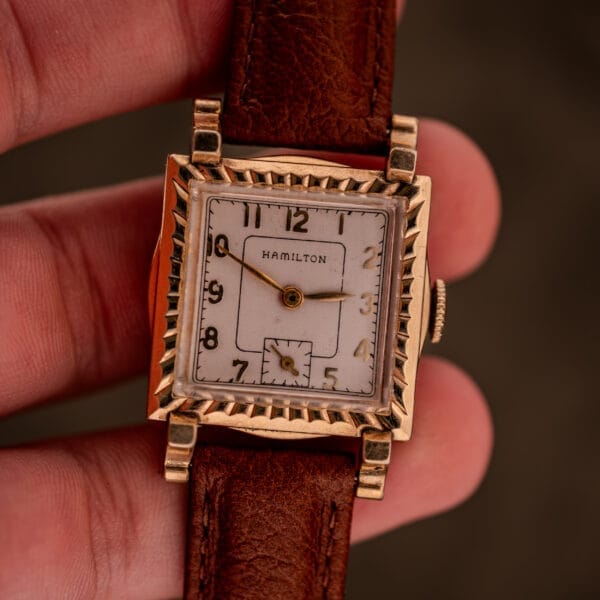 A hand holding a Hamilton Art Deco Square Gold Filled Case Manual Wind Cal. 987-F wristwatch with a vintage design and brown leather band.