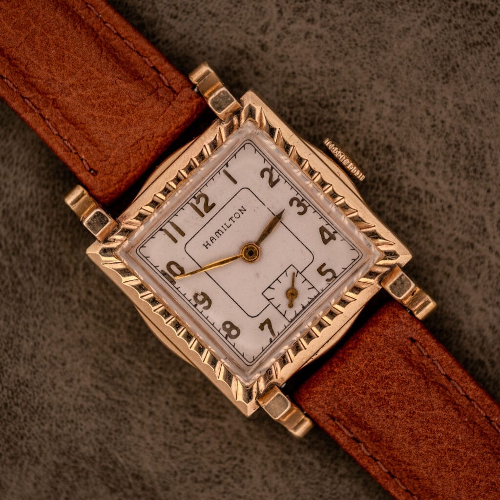 A Hamilton Art Deco Square Gold Filled Case Manual Wind Cal. 987-F wristwatch with a brown leather strap is displayed on a textured gray background.