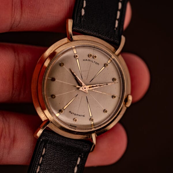A hand holds a Hamilton K-454 Sputnik Automatic Cal. 661 wristwatch with a gold filled case and black leather strap.