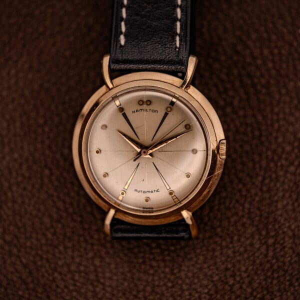 The Hamilton K-454 Sputnik Automatic Cal. 661 features a gold filled case and is shown with a cream dial and black leather strap on a brown textured surface.