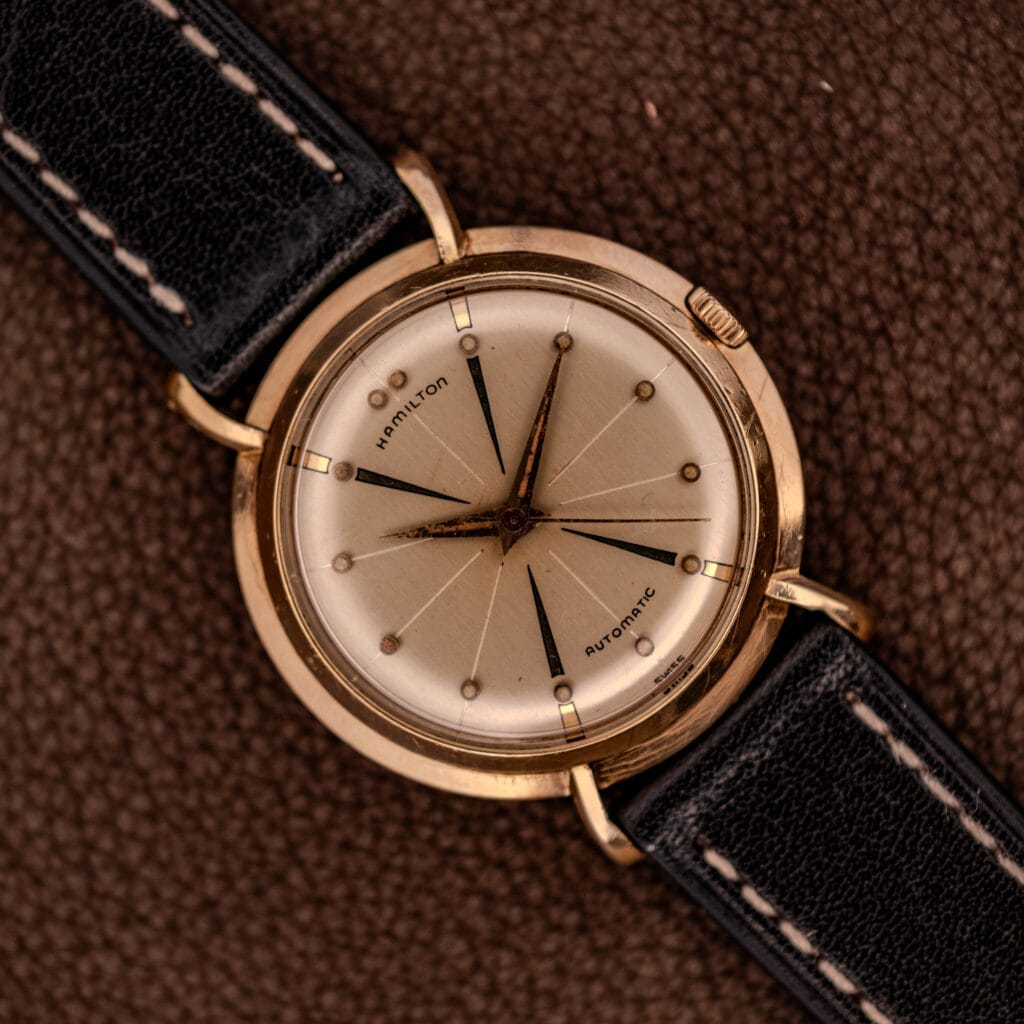 A Hamilton K-454 Sputnik Automatic Cal. 661 gold-filled wristwatch with a black leather strap is displayed elegantly on a brown textured surface.