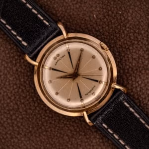 A Hamilton K-454 Sputnik Automatic Cal. 661 gold-filled wristwatch with a black leather strap is displayed elegantly on a brown textured surface.