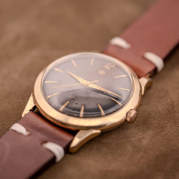 A Zodiac Hermetic Black Dial Automatic Caliber 1624 with a gold-filled case and brown leather strap rests elegantly on a soft brown surface.