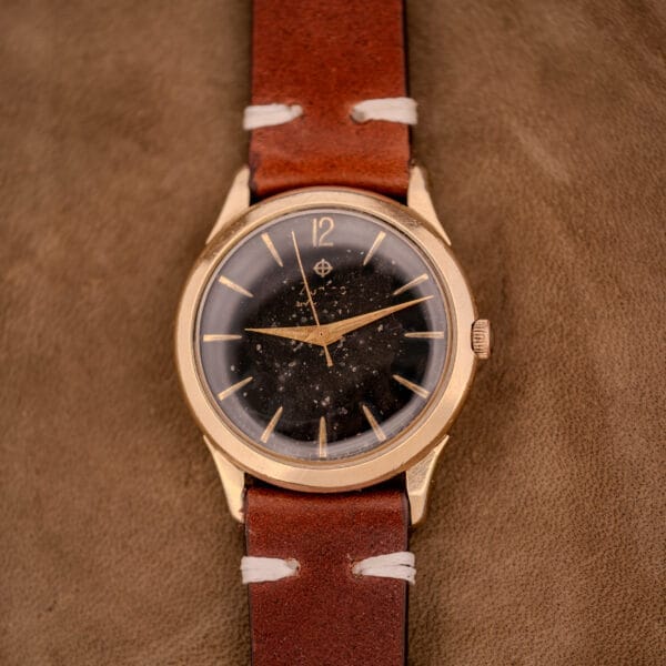 The Zodiac Hermetic Black Dial Automatic Caliber 1624 Gold Filled Case, with a brown leather strap and starry galaxy-themed dial, is showcased on a brown surface.