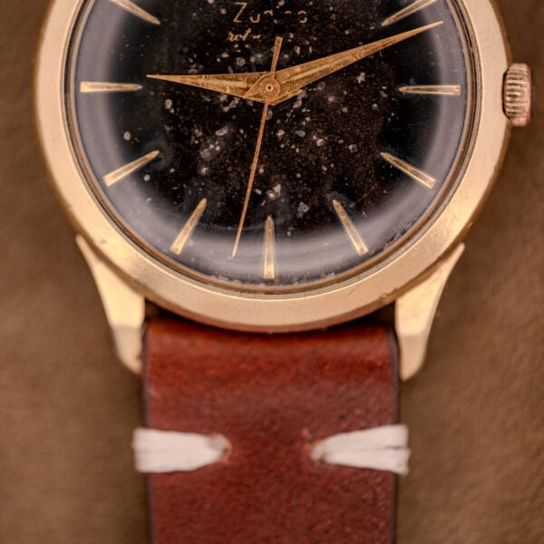 Close-up of a Zodiac Hermetic watch featuring a speckled black dial, gold filled case, brown leather strap, and Automatic Caliber 1624 movement.