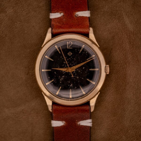 A Zodiac Hermetic Black Dial Automatic Caliber 1624 wristwatch with a Gold Filled Case and brown leather strap rests on a brown suede surface.