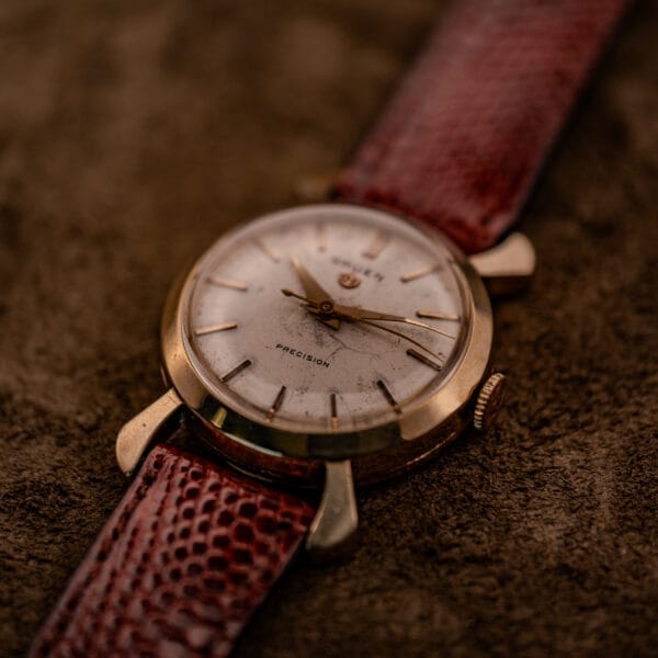 A Gruen 21 Precision Manual Wind Fancy Lugs Ref. 335SS 723 wristwatch with a brown leather strap rests on a soft brown fabric surface.