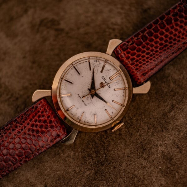 Gruen 21 Precision Manual Wind Fancy Lugs Ref. 335SS 723 wristwatch with gold case and brown textured leather strap, displayed on a brown surface; manual wind movement enhances its vintage appeal.