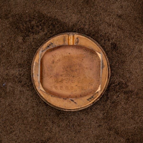 A worn, round, gold-toned metal piece with engraved numbers and text from a Gruen 21 Precision Manual Wind Fancy Lugs Ref. 335SS 723 watch, resting on brown textured fabric.