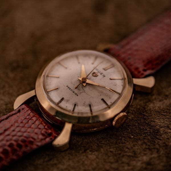 Close-up of a Gruen 21 Precision Manual Wind Fancy Lugs Ref. 335SS 723 gold wristwatch with a brown leather strap, resting on a textured brown surface.