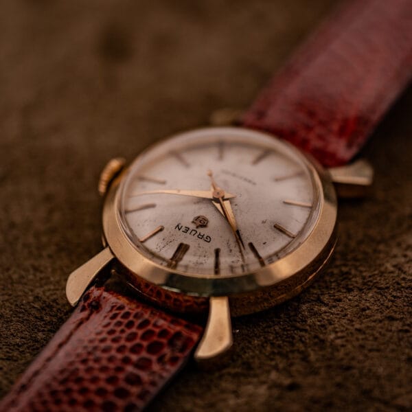 A Gruen 21 Precision Manual Wind Fancy Lugs Ref. 335SS 723 wristwatch with a brown leather strap is displayed on a textured brown surface.