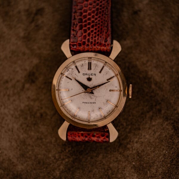 Gruen 21 Precision Manual Wind Fancy Lugs Ref. 335SS 723 wristwatch with a worn white dial and brown leather strap, displayed on a textured brown surface.