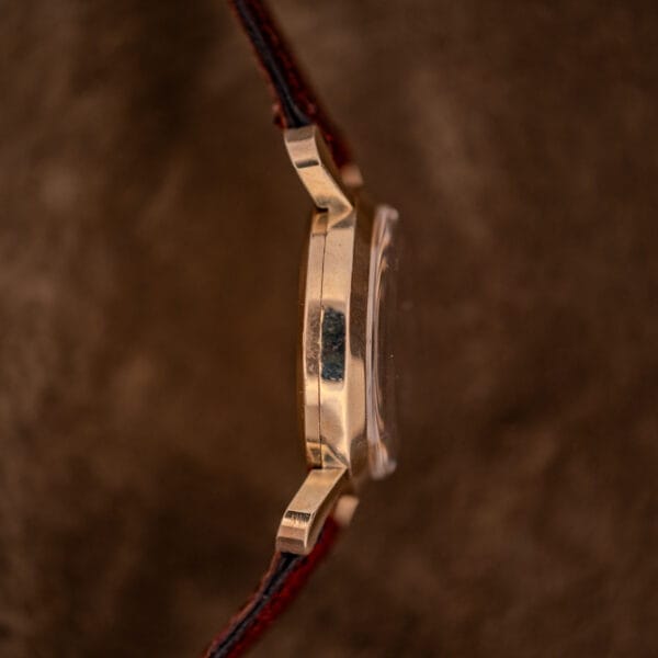 Side view of a Gruen 21 Precision Manual Wind Fancy Lugs Ref. 335SS 723 gold wristwatch with a brown leather strap, set against a textured brown background.