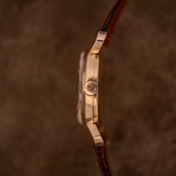 Side view of the Gruen 21 Precision Manual Wind Fancy Lugs Ref. 335SS 723 gold wristwatch with a brown leather strap, set against a soft brown textured background.