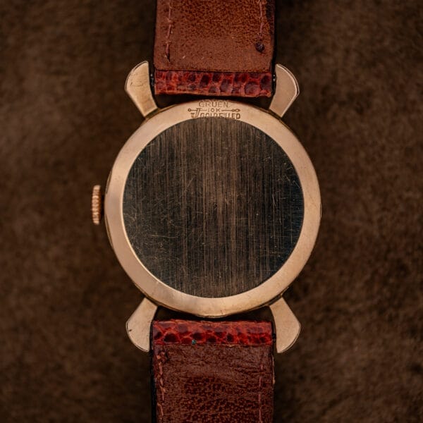 A Gruen 21 Precision Manual Wind Fancy Lugs Ref. 335SS 723 with a worn metal back and brown leather strap rests on a brown textured surface.