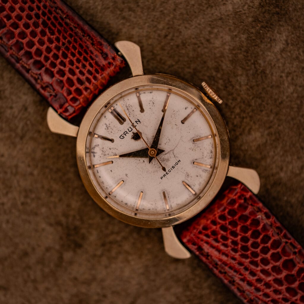 A Gruen 21 Precision Manual Wind Fancy Lugs Ref. 335SS 723 wristwatch with a gold case and red textured leather strap rests on a brown surface.