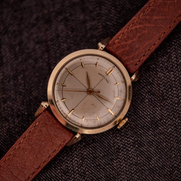 A Hamilton Rodney Sector Dial Manual Wind Cal. 748 wristwatch with a 10k gold filled case and brown leather strap rests on dark textured fabric.