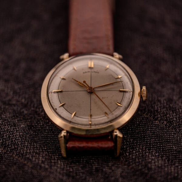 A Hamilton Rodney Sector Dial Manual Wind Cal. 748 wristwatch with a 10k gold filled case, shown with a brown leather strap on a textured dark fabric background.