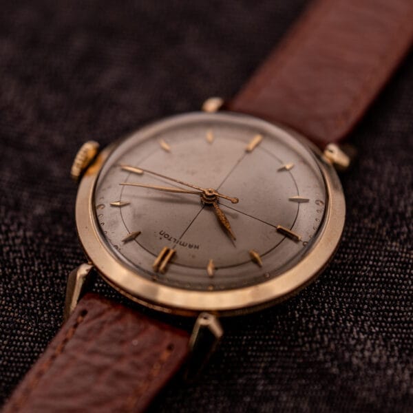 A Hamilton Rodney Sector Dial Manual Wind Cal. 748 wristwatch with a 10k gold filled case and brown leather strap rests on a dark textured surface.