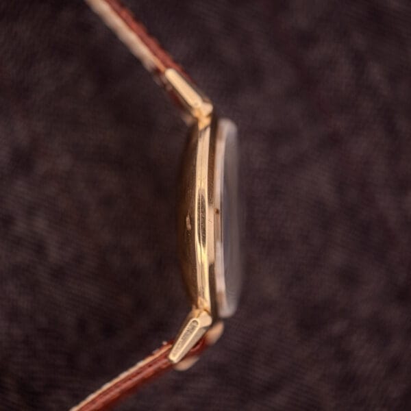 Side view of a Hamilton Rodney Sector Dial Manual Wind Cal. 748 wristwatch with a 10k gold filled case and brown leather strap, set on a textured dark background.