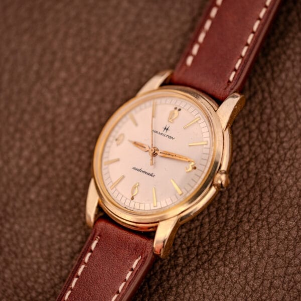 A Hamilton Automatic Cal. 689 wristwatch with a sunburst dial and 10k gold filled case, paired with a brown leather strap, rests on a textured brown surface.