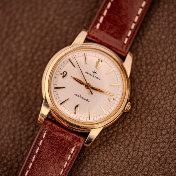 Hamilton Automatic Cal. 689 wristwatch with a sunburst dial, 10k gold filled case, and brown leather strap, displayed on a textured brown surface.