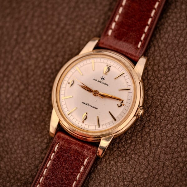 A Hamilton Automatic Cal. 689 Sunburst Dial 10k Gold Filled Case wristwatch with a brown leather strap rests on a textured brown surface.