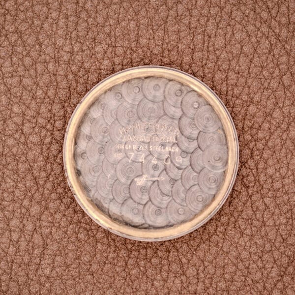 A silver case back with engraved text rests on textured brown leather—a subtle contrast to the Hamilton Automatic Cal. 689 Sunburst Dial's signature elegance and 10k gold filled case.