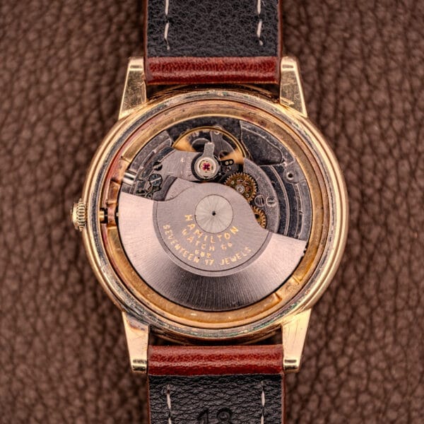 Close-up of a Hamilton Automatic Cal. 689 with sunburst dial and 10k gold filled case, showcasing its exposed mechanical movement on a brown textured surface.