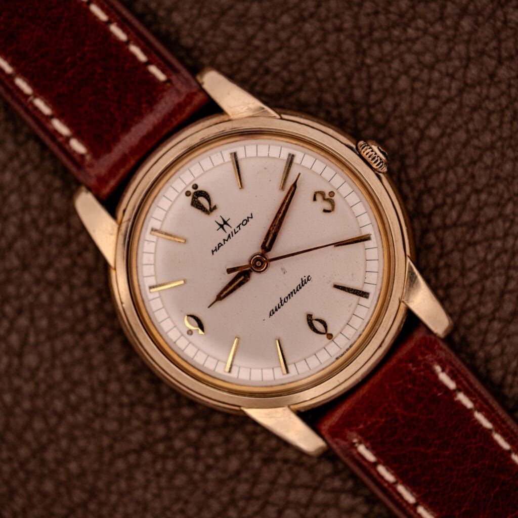 Hamilton Automatic Cal. 689 wristwatch with a sunburst dial and 10k gold filled case, paired with a brown leather strap, displayed on a textured brown surface.