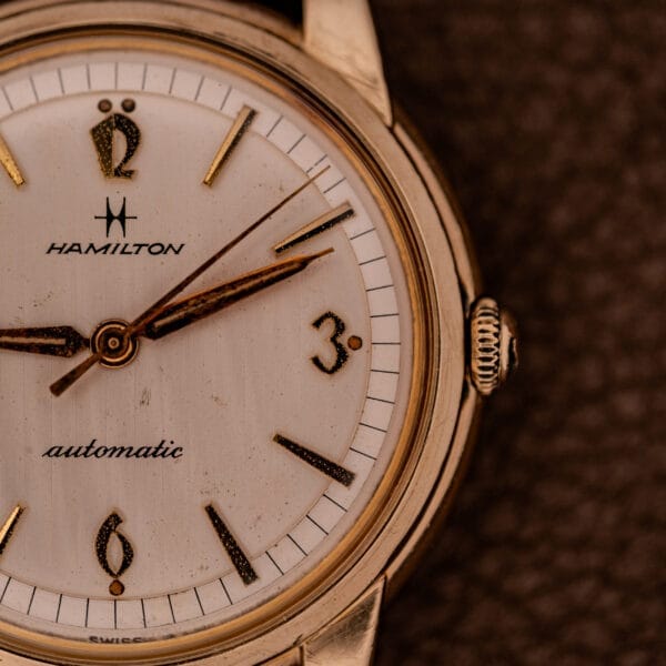 Close-up of a Hamilton Automatic Cal. 689 wristwatch featuring a sunburst dial and 10k gold filled case, displayed on a brown textured background.