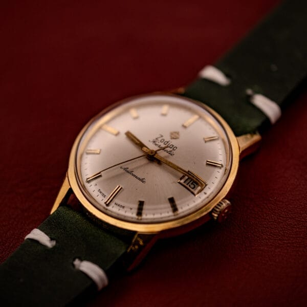 The Zodiac Hermetic Date Sunburst Dial Ref. 723-917 features a gold case, green leather strap, sunburst white dial with date display, and Automatic Cal. 70-72 movement.