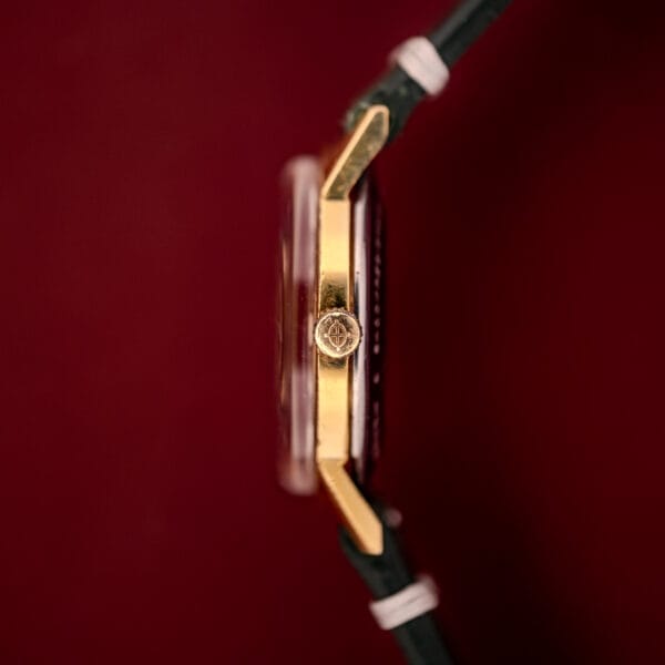 A close-up side view of the Zodiac Hermetic Date Sunburst Dial Ref. 723-917 Automatic Cal. 70-72 gold wristwatch with a black strap, showcased against a vivid red background.