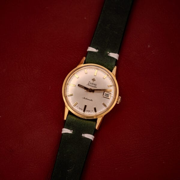 A Zodiac Hermetic Date Sunburst Dial Ref. 723-917 Automatic Cal. 70-72 wristwatch with a green leather band rests elegantly on a red leather surface.