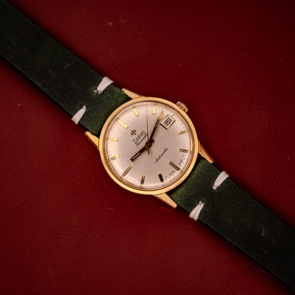 A Zodiac Hermetic Date Sunburst Dial Ref. 723-917 Automatic Cal. 70-72, featuring a gold case and green leather strap, rests elegantly on a dark red surface.