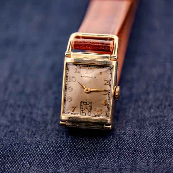 A Hamilton Willshire 14k Gold Filled Art Deco Case Cal. 980 wristwatch with a rectangular face and brown leather strap is displayed on blue fabric.