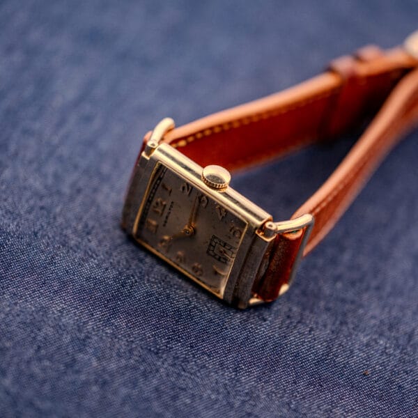 A Hamilton Willshire 14k Gold Filled Art Deco Case Cal. 980 wristwatch with a brown leather strap is displayed on blue fabric.