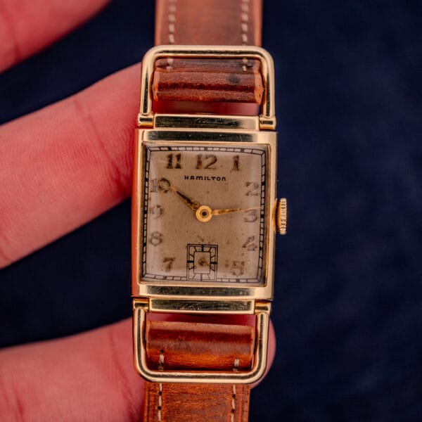A hand holds a Hamilton Willshire 14k Gold Filled Art Deco Case Cal. 980 watch with a rectangular case, brown leather strap, and classic Art Deco numeral dial.
