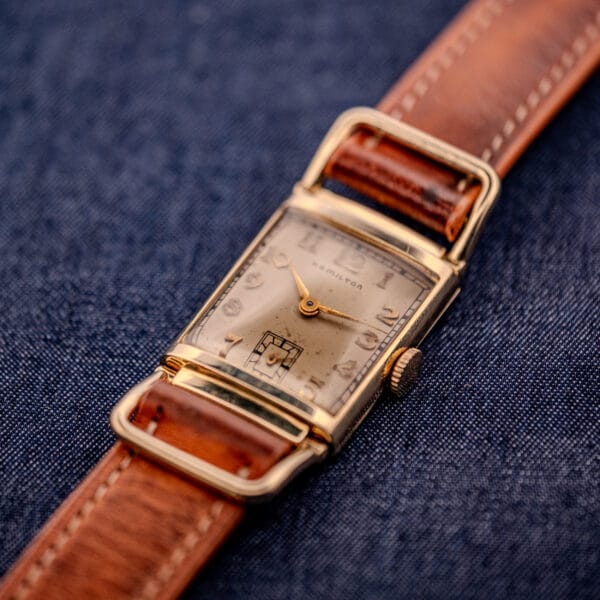A Hamilton Willshire 14k Gold Filled Art Deco Case Cal. 980 watch with a brown leather strap rests on blue denim fabric.