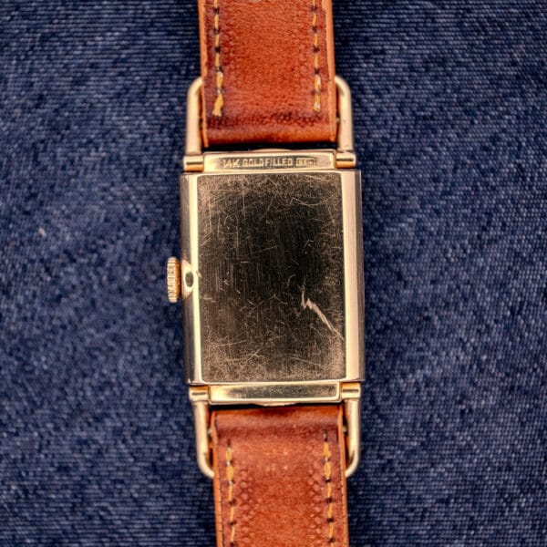 A Hamilton Willshire 14k Gold Filled Art Deco Case Cal. 980 wristwatch with a scratched back and brown leather strap is displayed on blue denim, highlighting its vintage elegance.