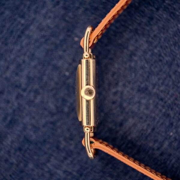 Side view of a Hamilton Willshire 14k Gold Filled Art Deco Case Cal. 980 wristwatch with a brown leather strap on a blue textured background.