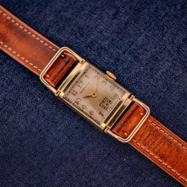 The Hamilton Willshire 14k Gold Filled Art Deco Case Cal. 980 wristwatch with a brown leather strap is displayed on dark blue fabric, embodying timeless Art Deco elegance.