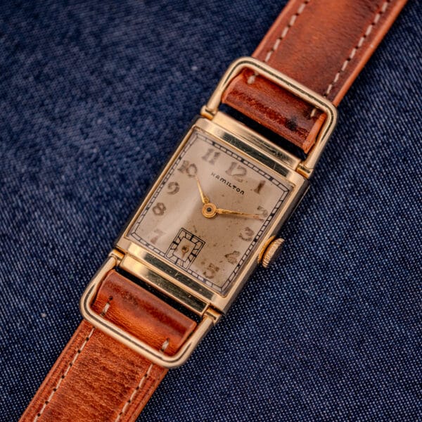 Hamilton Willshire 14k Gold Filled Art Deco Case Cal. 980 wristwatch with a rectangular case and brown leather strap, shown on blue denim fabric.