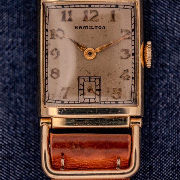 Close-up of a Hamilton Willshire 14k Gold Filled Art Deco Case Cal. 980 watch with a brown leather band, displayed on blue fabric.