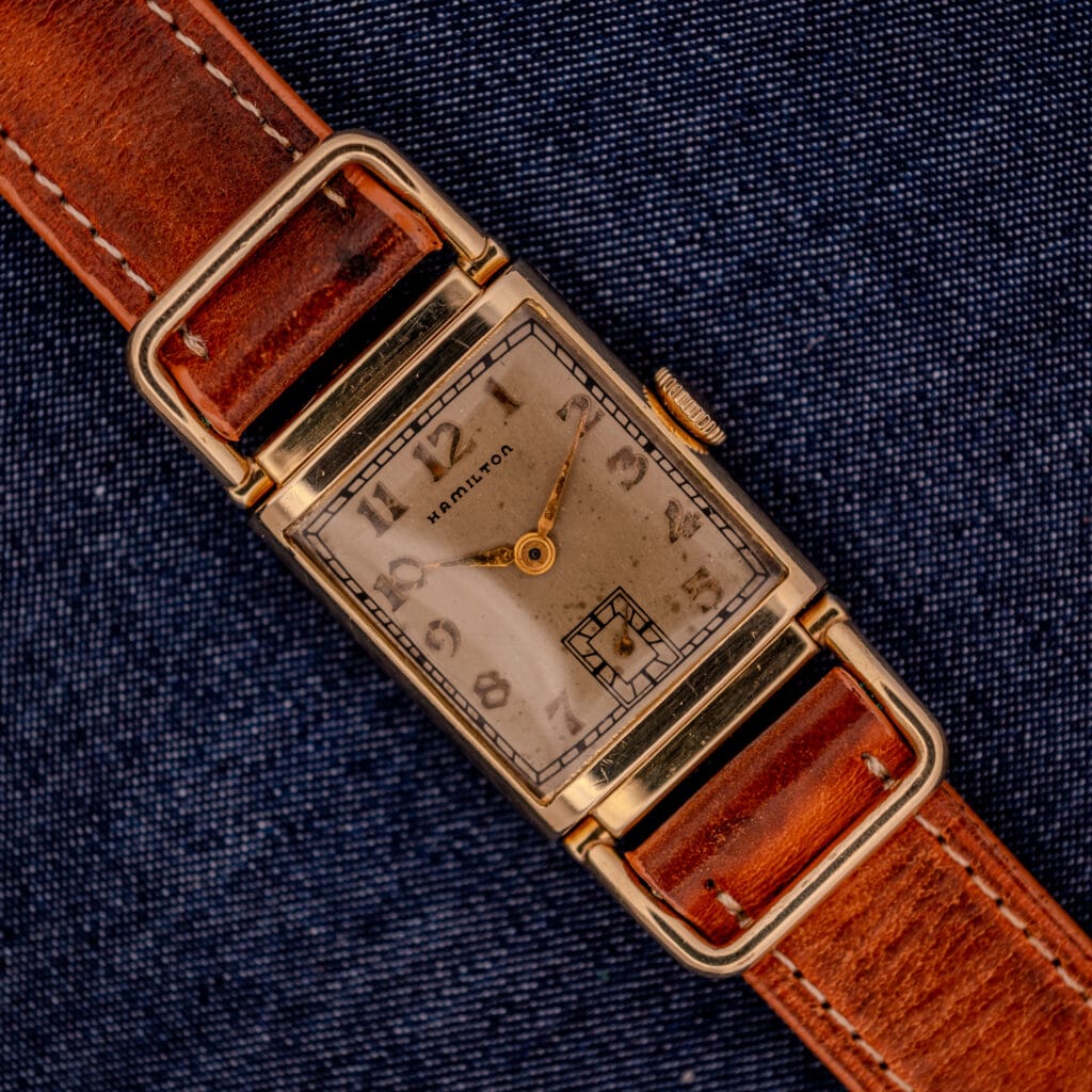 A Hamilton Willshire 14k Gold Filled Art Deco Case Cal. 980 watch with a brown leather strap rests on dark blue fabric.