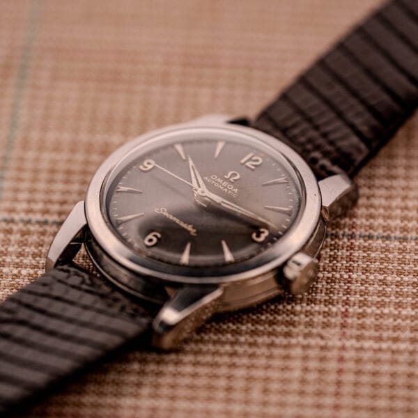 A 1956 Omega Seamaster Automatic Ref. 2828-6 SC Cal. 470 Stainless Steel wristwatch with a black dial and leather strap rests elegantly on a plaid surface.