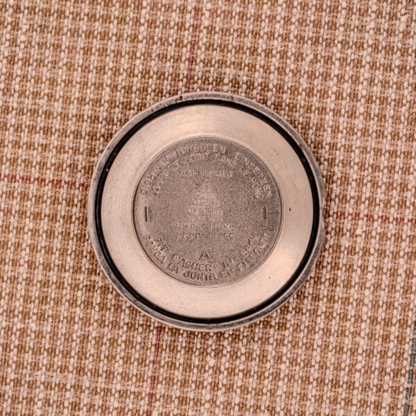 A round metallic lid with embossed text and symbols, likely from a 1956 Omega Seamaster Automatic Ref. 2828-6 SC Cal. 470 Stainless Steel, rests on a brown plaid fabric surface.