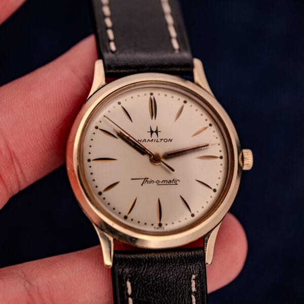 A hand holds a 1962 Hamilton Thin-o-Matic Cal. 663 10K Gold Filled wristwatch, featuring a black leather strap and cream dial.