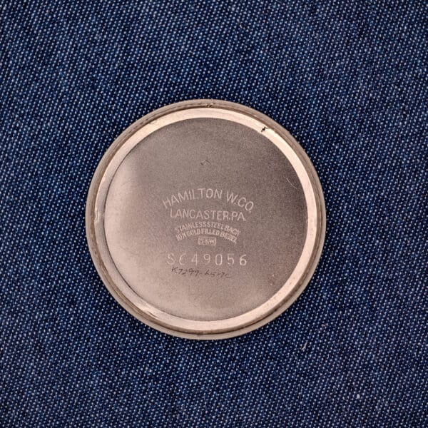 A round metal case back on blue fabric, engraved with Hamilton W.Co Lancaster, PA, Cal. 663, and serial numbers, from a 1962 Hamilton Thin-o-Matic Cal. 663 10K Gold Filled model.