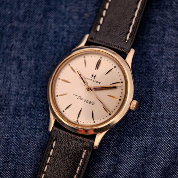 The 1962 Hamilton Thin-o-Matic Cal. 663 10K Gold Filled wristwatch with a black leather strap is displayed on a blue denim fabric background.